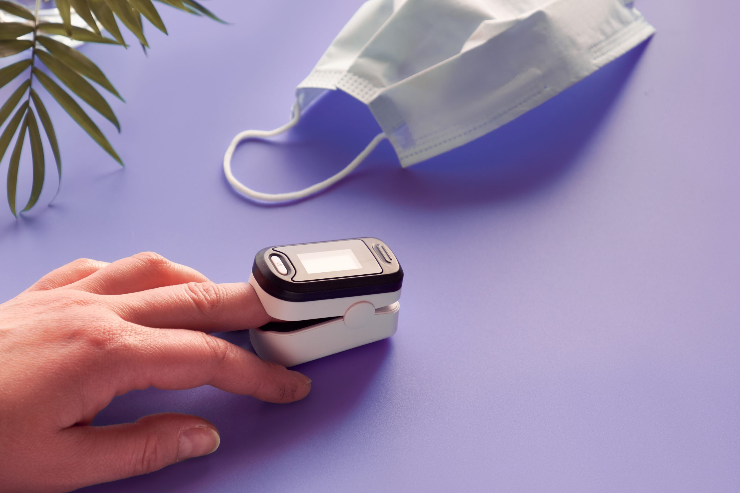 pulse-oximeter-device-monitoring-oxygen-saturation-reduced-oxygenation-is-emergency-sign-covid-19-viral-pneumonia-digital-device-female-finger-face-mask-palm-plant-purple-neon-table-scaled