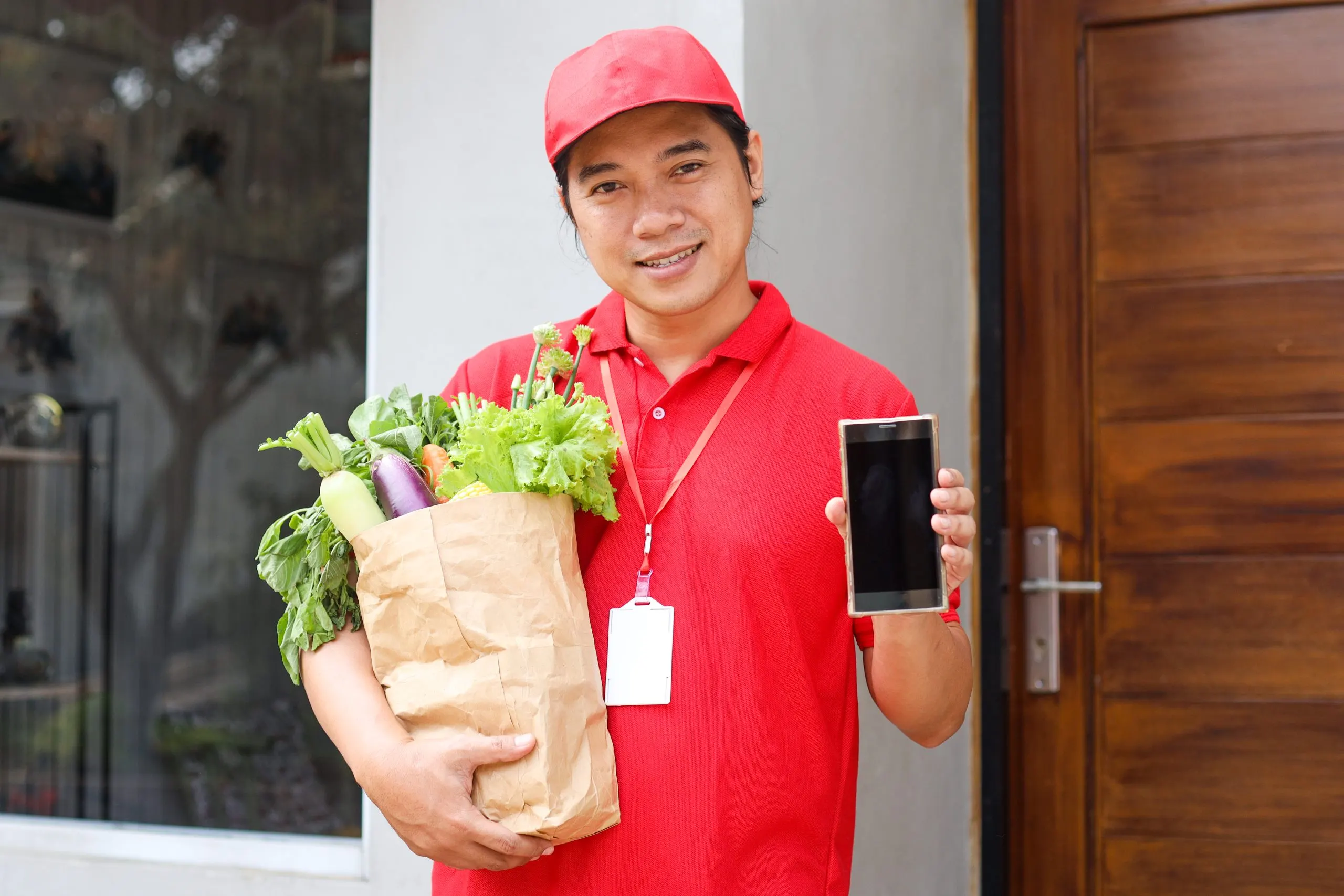 Philippines Online Grocery Delivery Market Outlook to 2035 asian-deliveryman-holding-paper-bag-with-food-vegetables-showing-blank-phone-screen-scaled