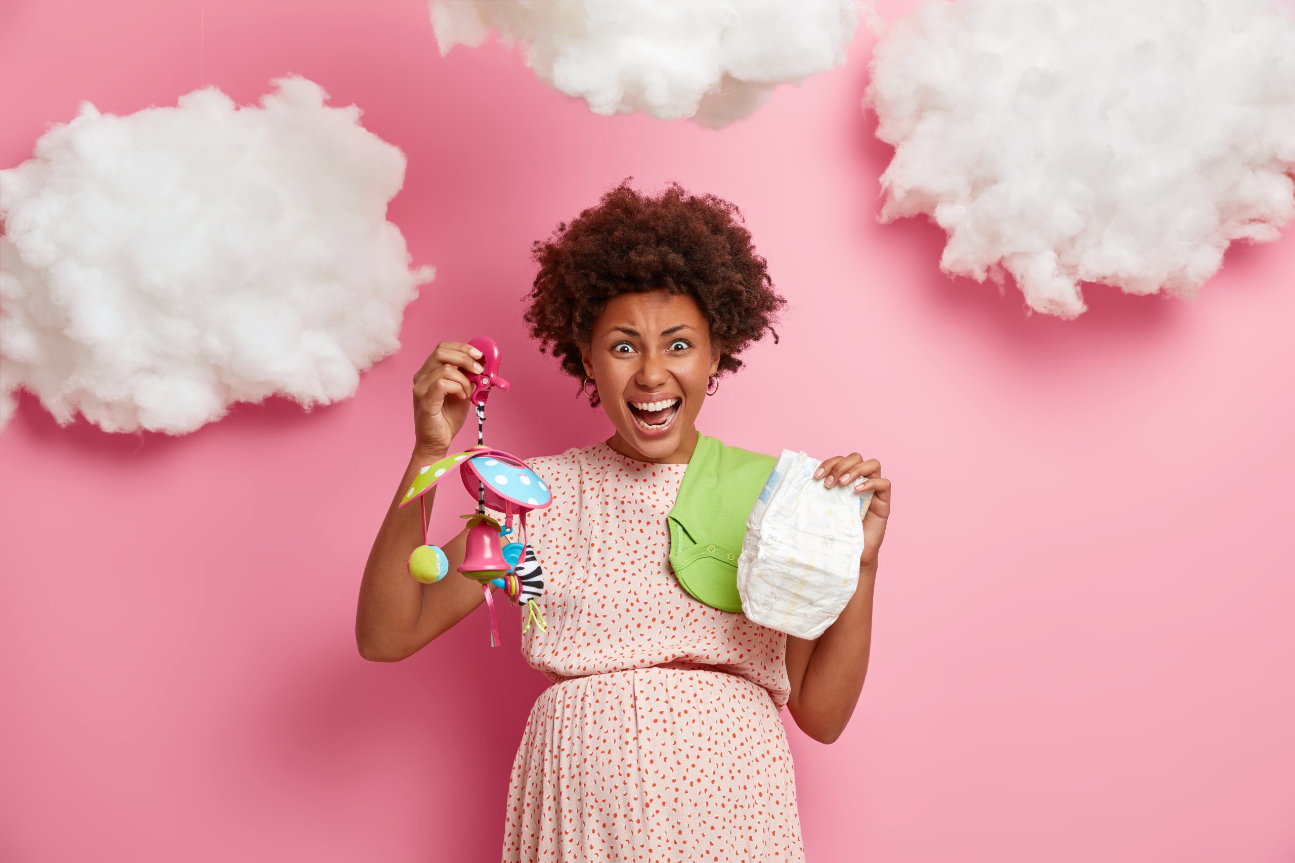 emotional-expectant-mother-shouts-emotionally-holds-diaper-baby-mobile-prepares-child-birth-wears-dress-prepares-baby-shower-poses-against-pink-wall-white-clouds-overhead-scaled