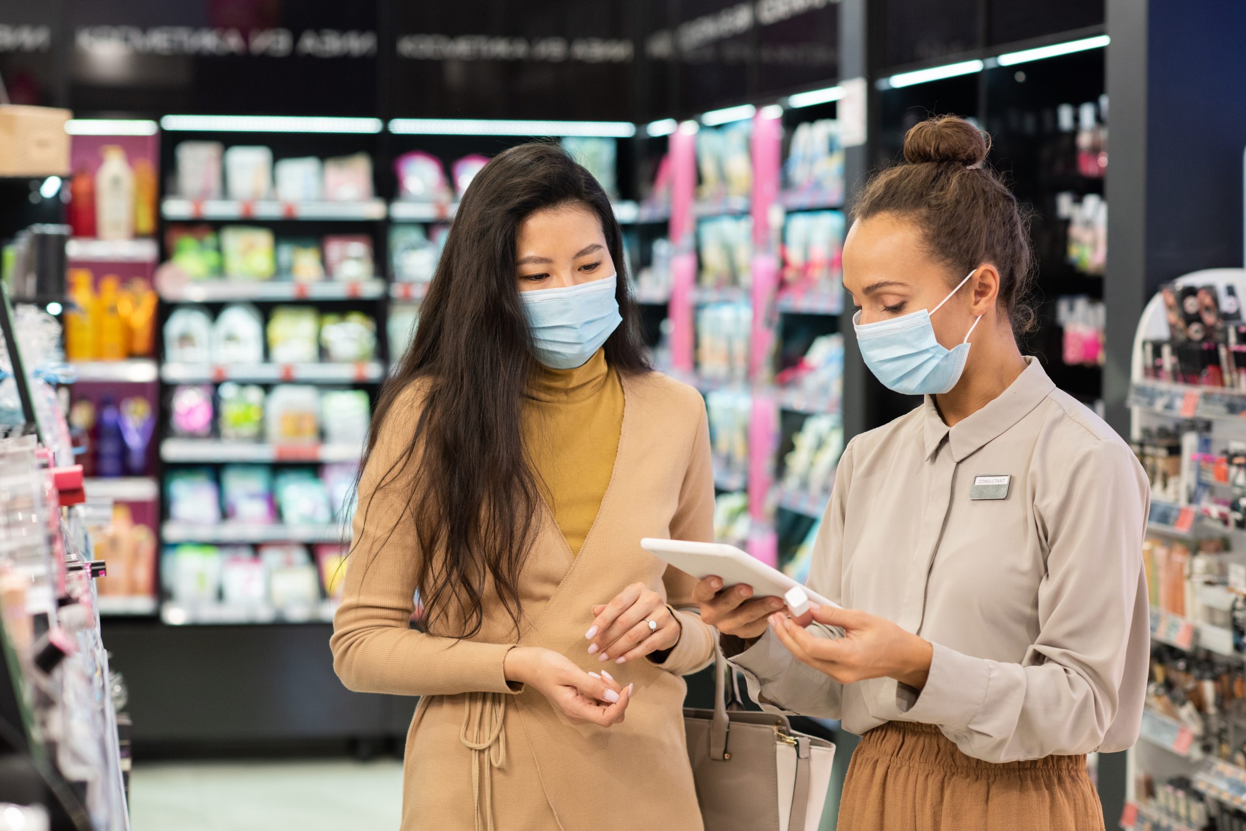 young-consultant-protective-mask-scrolling-tablet-while-looking-price-lip-gloss-wanted-by-one-clients-beauty-supermarket-scaled