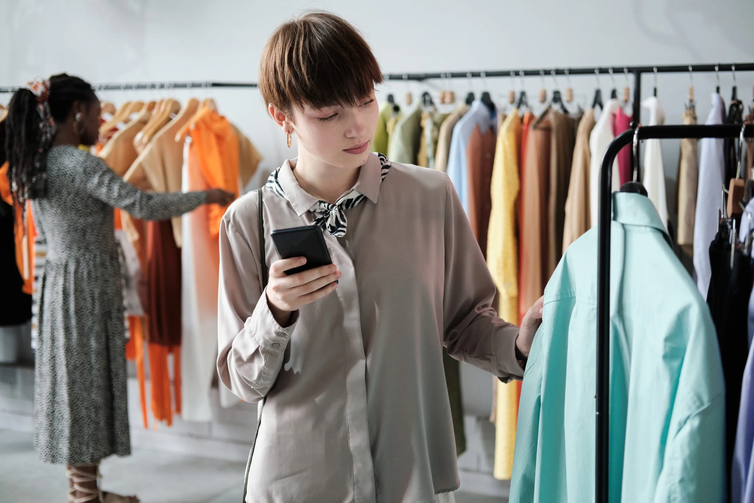 UK Online Clothing Rental Market Outlook to 2035 young-woman-choosing-new-shirts-rack-while-using-her-mobile-phone-shopping-mall-scaled