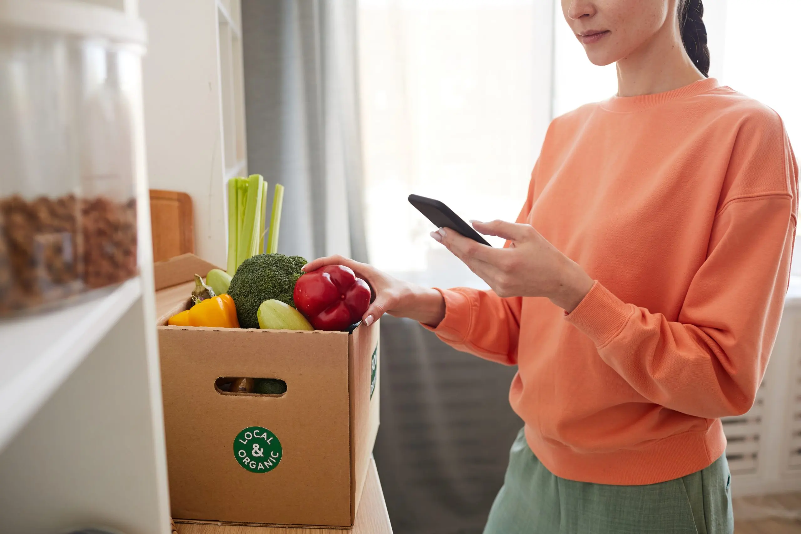 UK Online Grocery Delivery Market Expands as 20 Percent of Consumers Shop Food Online close-up-young-woman-typing-message-her-mobile-phone-while-standing-near-cardboard-box-with-fresh-vegetables-scaled