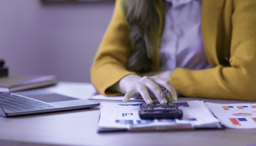 Close-up of a corporate finance manager using an electronic calculator over printed charts, financial statements, and a laptop on an office desk.
